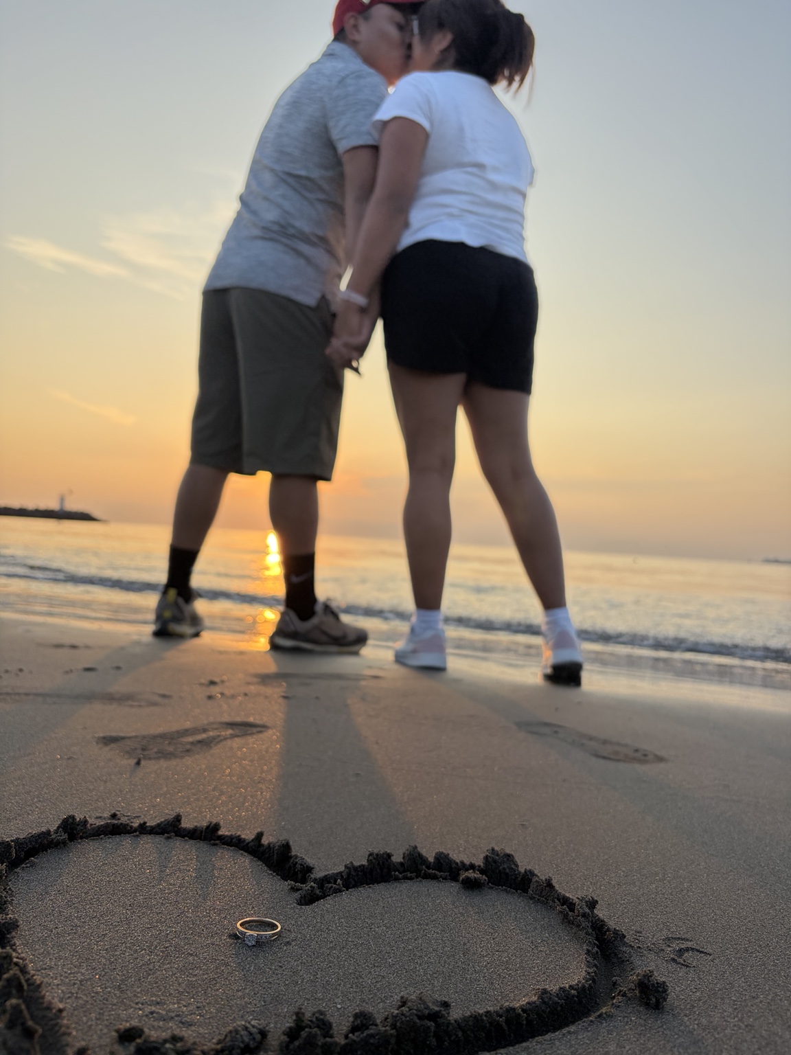 Felipe y Belén en la playa al atardecer, con un corazón y un anillo en la arena